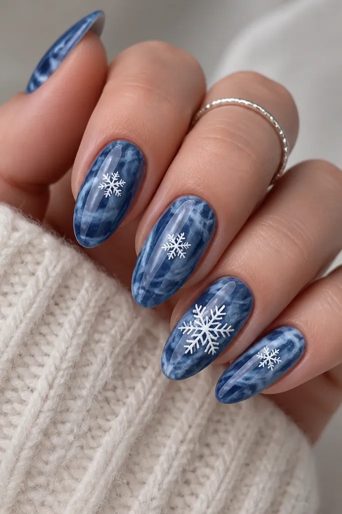Medium almond nails with a denim-blue marbled swirl background. White snowflake decals sit near the center of each nail, with one larger snowflake on the ring finger. A glossy top coat reflects light in smooth highlights.