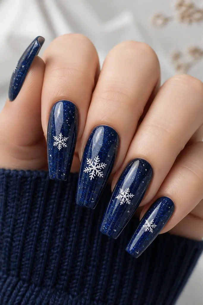 Long coffin nails in deep navy. Each nail has a faint "star" gradient using tiny silver foil specks. A single white snowflake sits mid-nail on the index and ring fingers, with smaller ones on the others. Highly glossy finish.