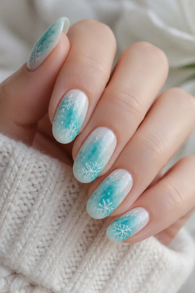 Medium oval nails with aqua watercolor swirls that bleed into each other over a milky white base. White snowflakes are painted loosely, with small white splatter dots around them. Glossy top coat makes the watercolor look wet.