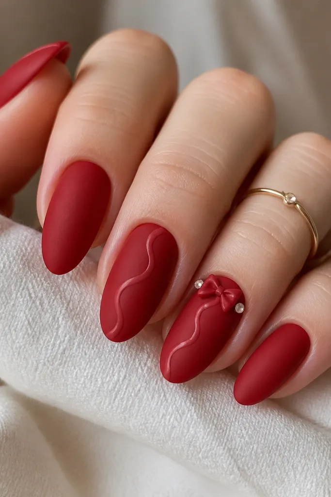 All nails are matte velvet red. Two nails have a thin glossy swirl in a lighter red near the center. On one accent nail, there is a tiny 3D bow charm at the cuticle with two micro rhinestones as 'lights' on each side.