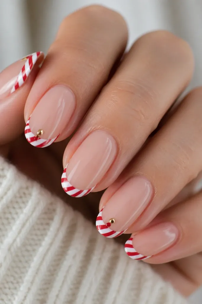 Most nails are glossy sheer nude. French tips are candy cane stripes - alternating red and white diagonal bands. Two nails have a tiny gold plug-like rectangle at the base of the tip, and a small red dot sits under it like a bulb.